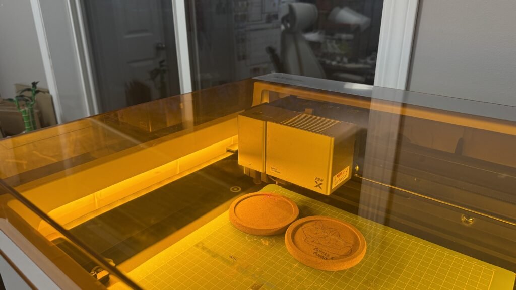 xTool M1 Ultra laser engraving machine engraving custom cork coasters with Coffee & Cookie cat design inside a warm-lit creative studio workspace.