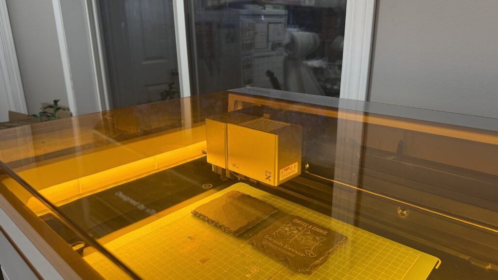Behind-the-scenes photograph of a laser engraving machine actively engraving a personalized square slate pet memorial coaster inside the machine enclosure with warm amber lighting.