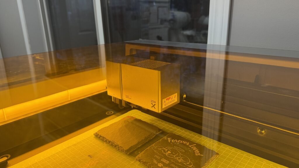 Laser engraving machine engraving a custom pet memorial slate coaster inside a warmly lit workshop.
