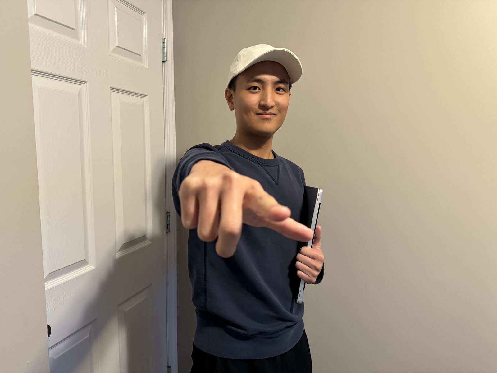 Evan is smiling and wearing a white cap pointing toward the camera while holding a laptop indoors.