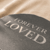 Close-up photograph of a round black slate memorial coaster engraved with the words “Forever Loved,” resting on softly textured cream linen fabric in warm natural sunlight.