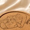Close-up angled product photo of a personalized cork coaster engraved with two orange tabby cats cuddling together and the text “Coffee & Cookie,” displayed on soft cream linen fabric under warm natural lighting.