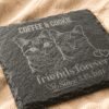 Close-up angled view of a personalized engraved square slate pet memorial coaster featuring custom cat portrait artwork and family name engraving on soft cream linen fabric.