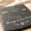 Close-Up of Personalized Slate Memorial Coaster Close-up angled photograph of a personalized square slate pet memorial coaster engraved with custom pet portraits, names, memorial year, and “Always in Our Hearts” text on a soft linen fabric background.