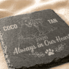 Close-up angled photograph of a personalized square slate pet memorial coaster engraved with custom pet portraits, names, memorial year, and “Always in Our Hearts” text on a soft linen fabric background.