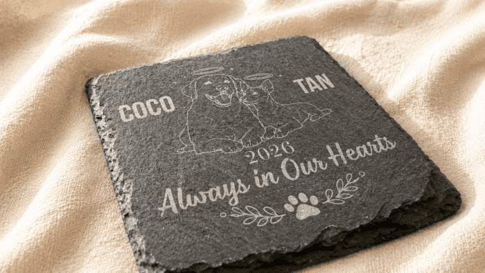 Close-up angled photograph of a personalized square slate pet memorial coaster engraved with custom pet portraits, names, memorial year, and “Always in Our Hearts” text on a soft linen fabric background.