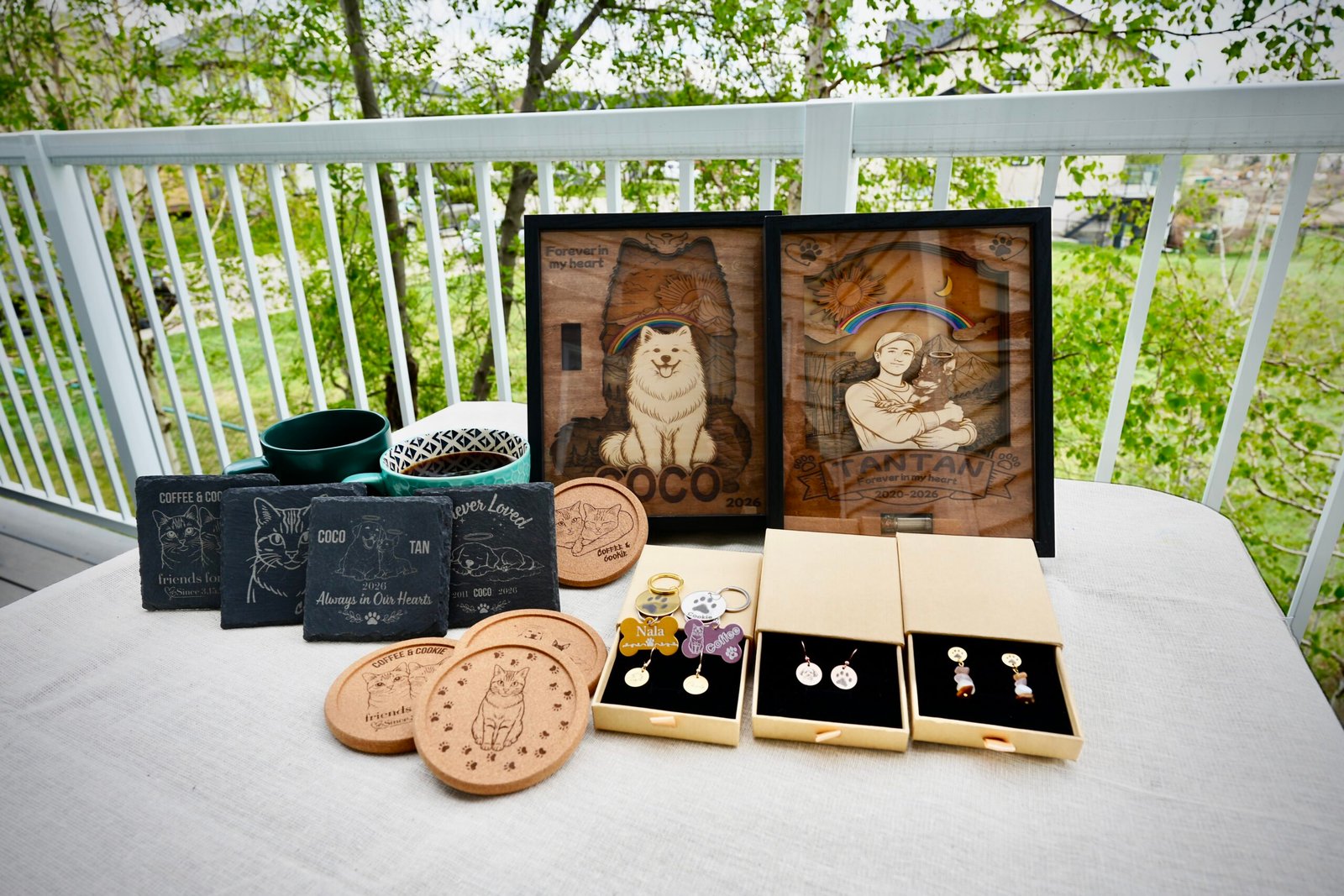 Collection of handcrafted personalized pet memorial gifts displayed on an outdoor table, including wooden shadow boxes, engraved slate coasters, cork coasters, pet tags, and memorial jewelry.