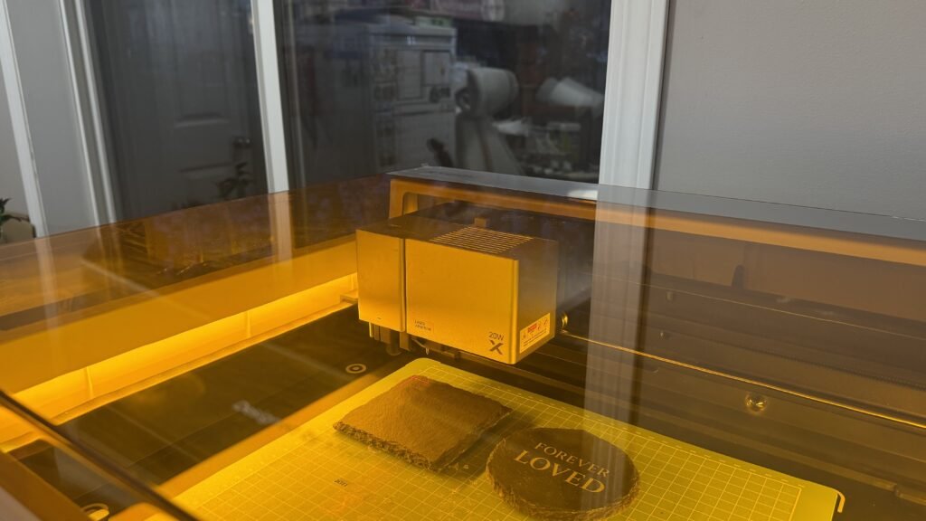 Behind-the-scenes photograph of an xTool 20W laser engraving machine engraving slate memorial coasters inside a softly lit workshop with warm amber reflections.