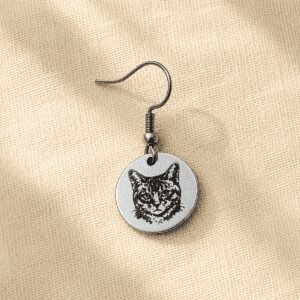 Personalized silver cat portrait earring featuring a custom engraved cat illustration on a round stainless steel charm, displayed on soft cream linen fabric in warm natural light.
