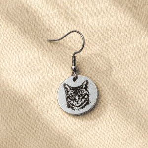 Personalized silver cat portrait earring featuring a custom engraved cat illustration on a round stainless steel charm, displayed on soft cream linen fabric in warm natural light.