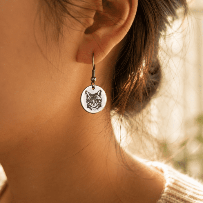Close-up lifestyle photo of a woman wearing a silver engraved cat portrait earring in warm golden natural light with soft blurred background.