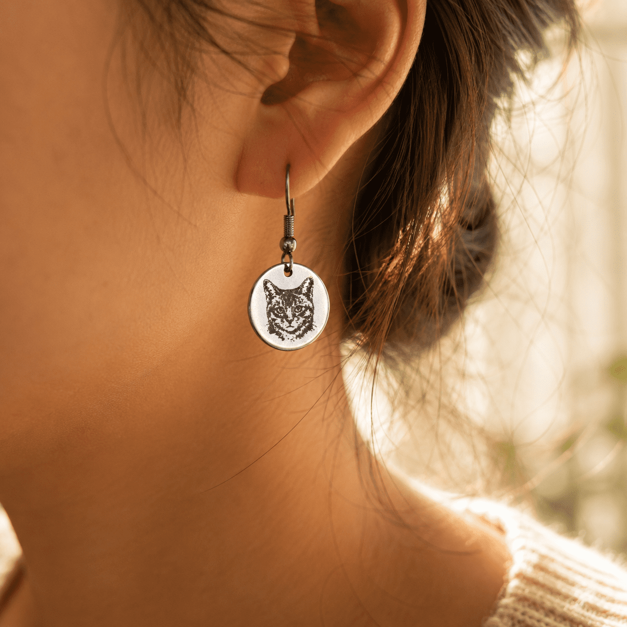 Close-up lifestyle photo of a woman wearing a silver engraved cat portrait earring in warm golden natural light with soft blurred background.