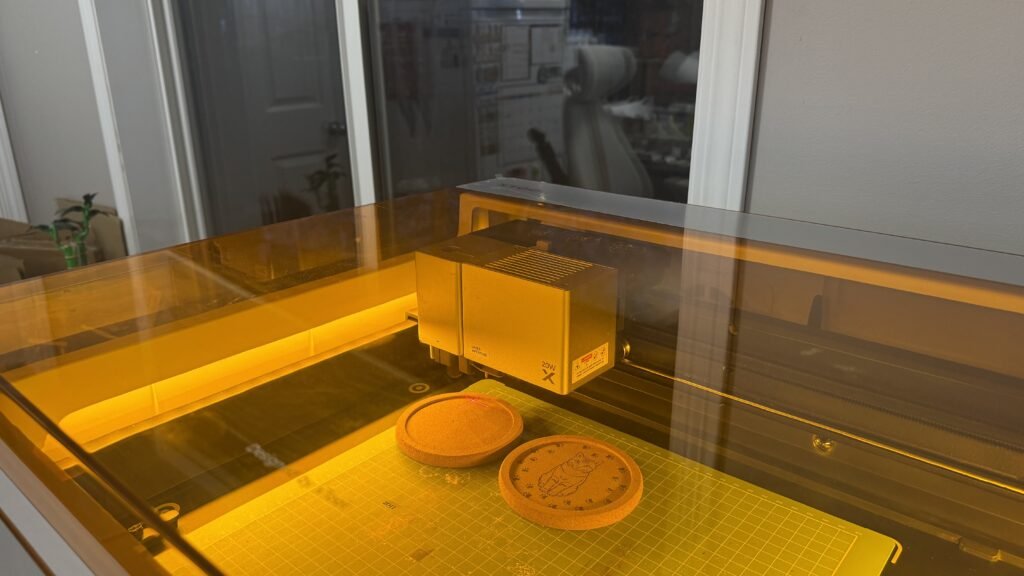 Laser engraving machine creating custom cork pet coasters with engraved cat artwork inside a home workshop studio.