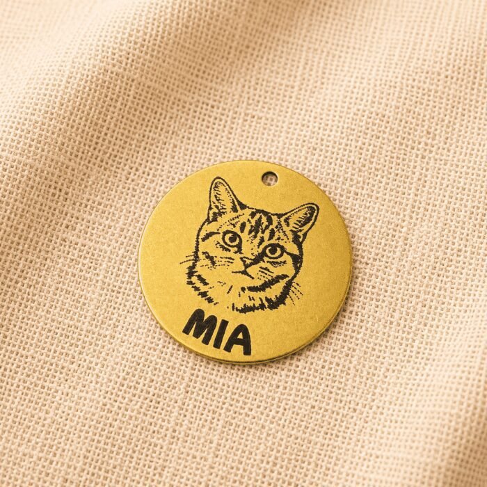 Close-up photograph of a personalized gold-tone pet charm engraved with a cat portrait and the name “MIA,” displayed on soft cream linen fabric.