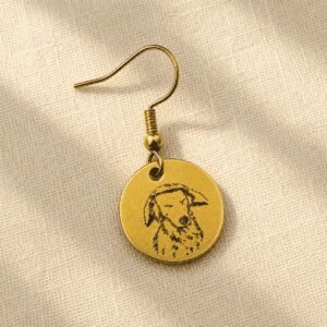 Personalized gold-tone pet memorial earring featuring a custom engraved dog portrait on a round charm, displayed on soft beige linen fabric in warm natural light.