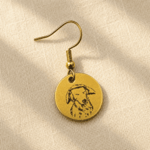 Personalized gold-tone pet memorial earring featuring a custom engraved dog portrait on a round charm, displayed on soft beige linen fabric in warm natural light.