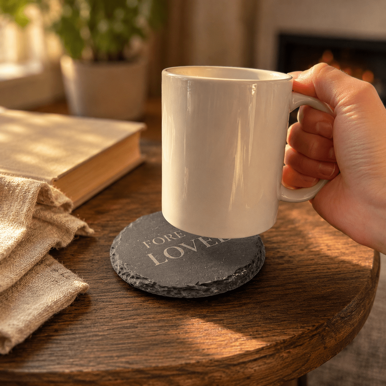 Hand holding a white ceramic mug above a round black slate memorial coaster engraved with “Forever Loved,” placed on a rustic wooden table in a warm cozy home setting.