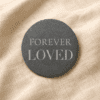 Minimalist round black slate memorial coaster engraved with the words “Forever Loved,” displayed on soft cream linen fabric with natural warm lighting.