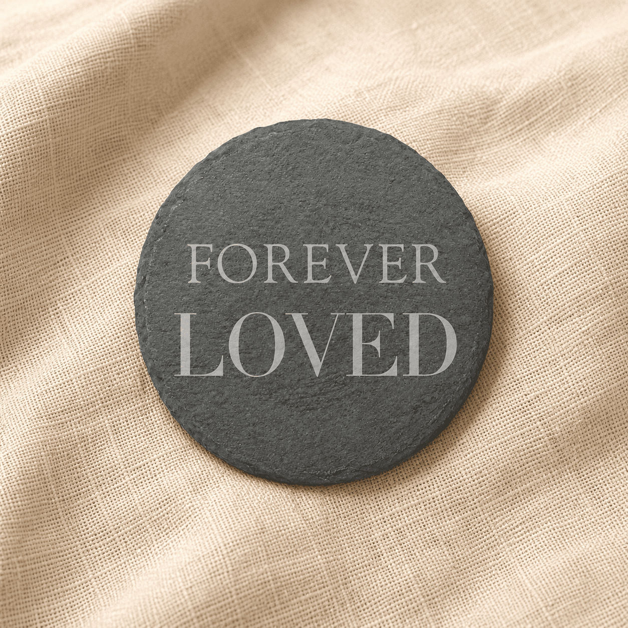 Minimalist round black slate memorial coaster engraved with the words “Forever Loved,” displayed on soft cream linen fabric with natural warm lighting.