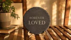 Round black slate memorial plaque engraved with the words “Forever Loved,” displayed on a wooden easel stand in a warm sunlit home setting with soft golden shadows.