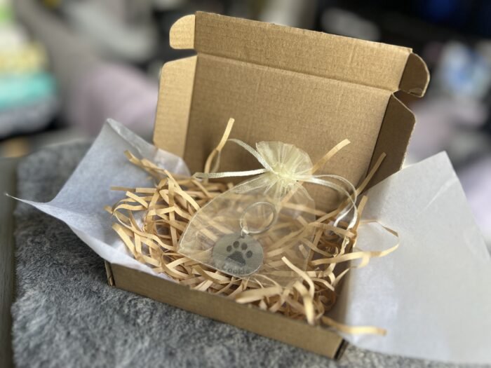 Open kraft gift box with tissue paper, shredded paper filler, and a personalized paw print keychain packaged inside a soft organza pouch.