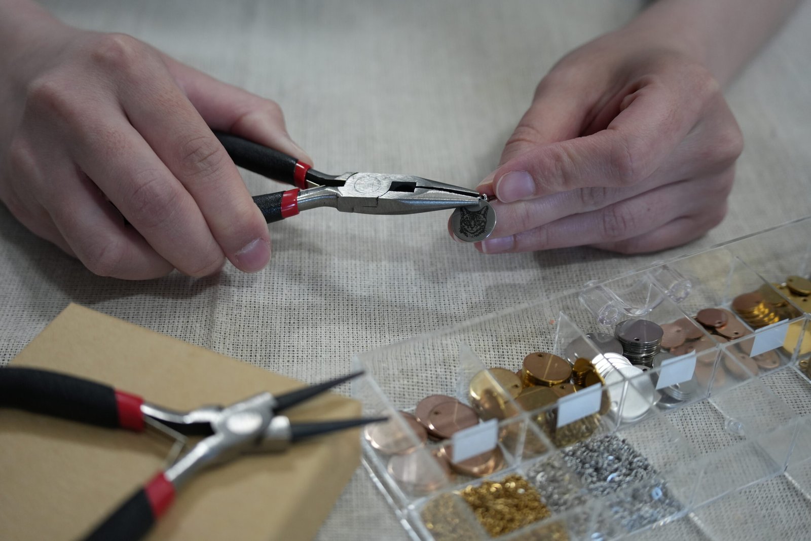 Hands carefully assembling a personalized engraved pet charm using jewelry pliers, surrounded by metal blanks, findings, and crafting tools on a neutral linen workspace.
