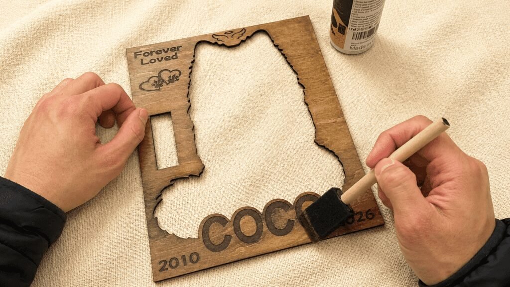 Close-up overhead photo of a handcrafted wooden pet memorial shadow box layer being hand-finished with walnut stain using a foam brush on a linen surface.