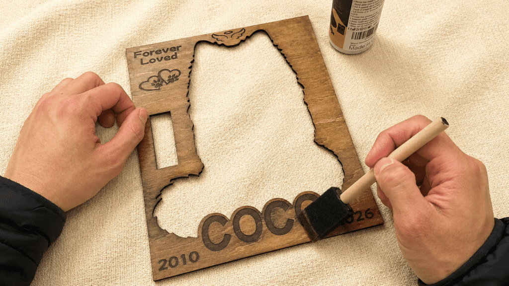 Close-up overhead photo of a handcrafted wooden pet memorial shadow box layer being hand-finished with walnut stain using a foam brush on a linen surface.