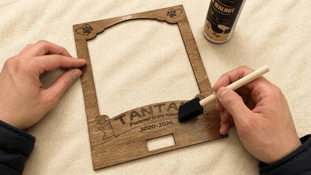Hands applying walnut finish to a laser engraved wooden pet memorial shadow box frame using a foam brush during the handmade crafting process.