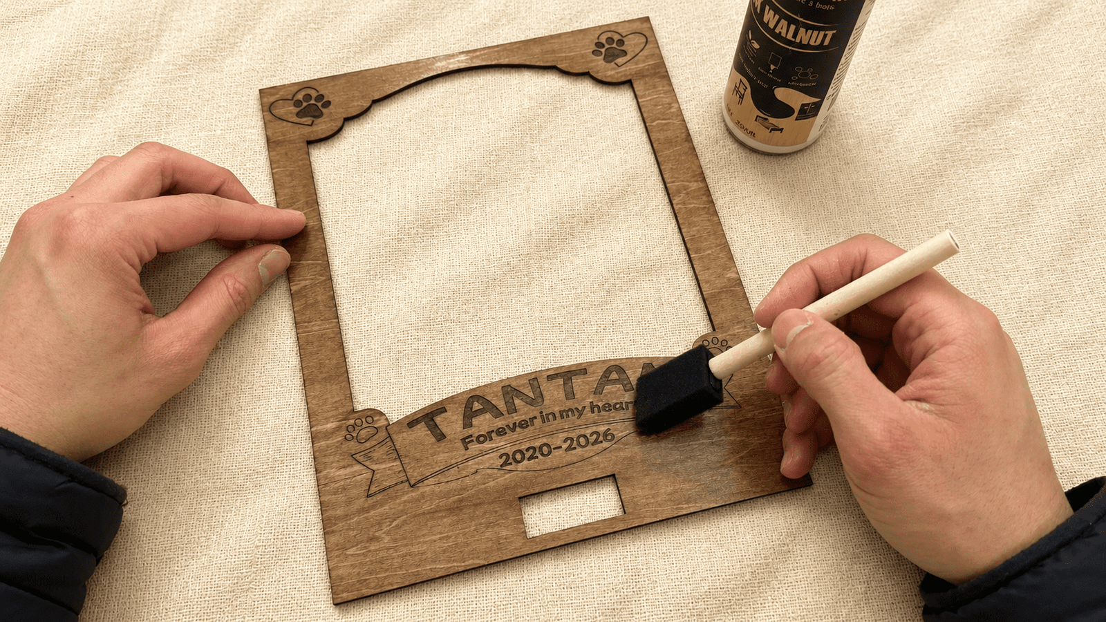 Hands carefully assembling a personalized engraved pet charm using jewelry pliers, surrounded by metal blanks, findings, and crafting tools on a neutral linen workspace.