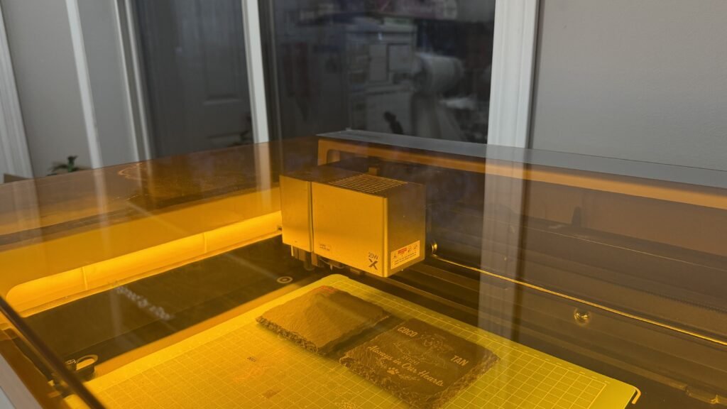 Behind-the-scenes photo of a laser engraving machine engraving personalized slate memorial coasters inside an xTool M1 Ultra workspace with warm amber lighting.