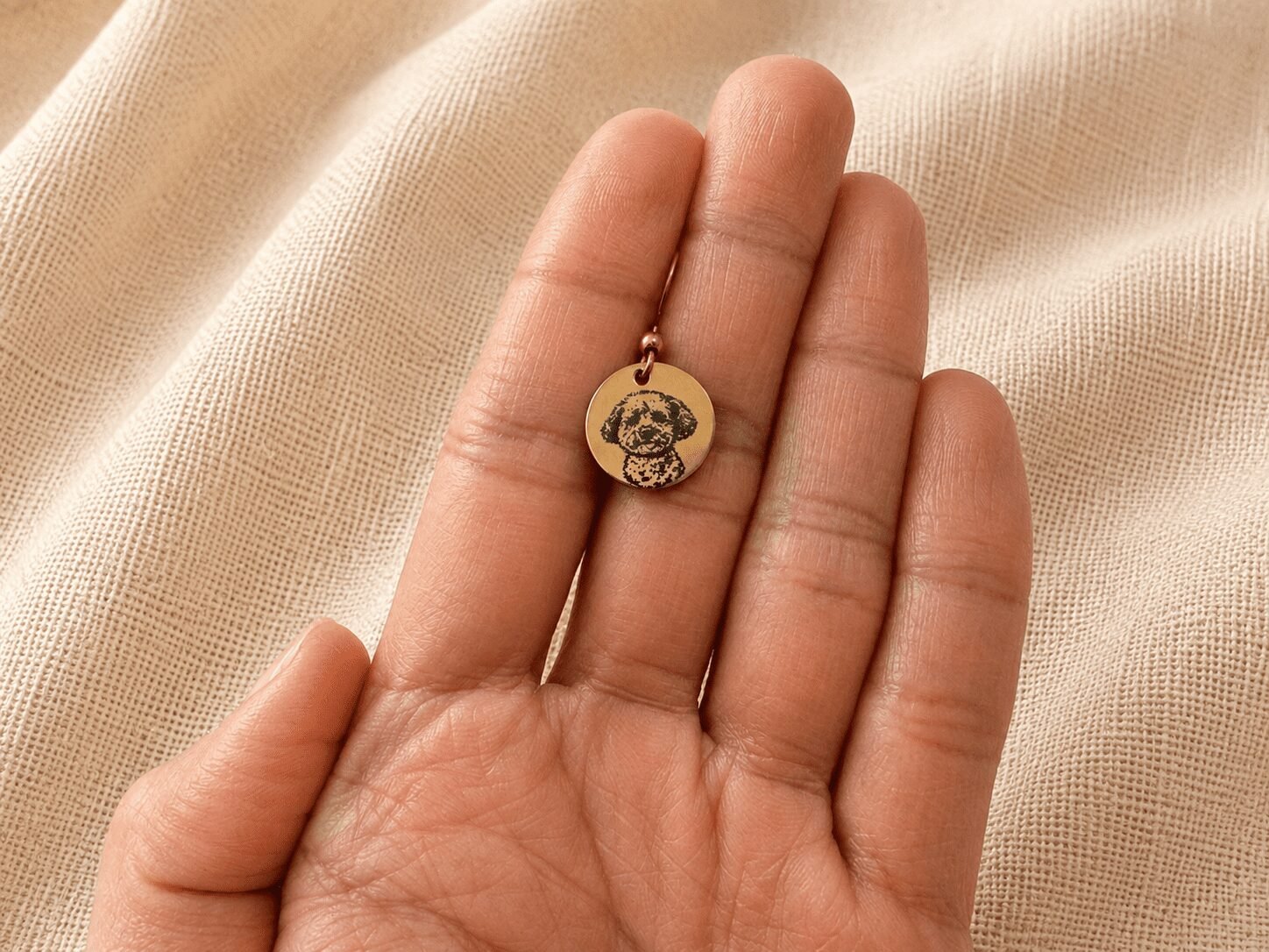 Close-up of a hand holding a tiny rose gold engraved pet charm with a custom dog portrait, shown against soft beige linen fabric for size reference.