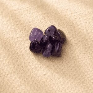 Polished amethyst crystal chips with rich purple hues displayed on a neutral beige linen fabric background in soft natural lighting.