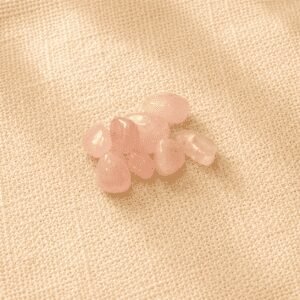 Polished rose quartz crystal chips with soft blush pink tones displayed on a beige linen fabric background under natural light.