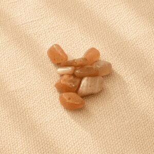 Polished sunstone crystal chips with warm peach and golden-orange tones arranged on a beige linen fabric background under soft natural light.
