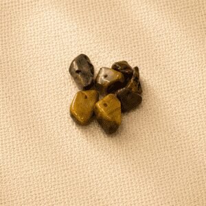 Polished tiger’s eye crystal chips with golden brown and dark amber banding displayed on a beige linen fabric background in soft natural light.