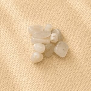 Polished white moonstone crystal chips with soft translucent white tones displayed on a beige linen fabric background in natural light.