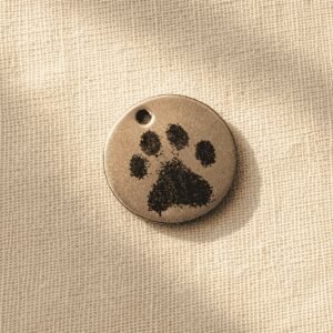 Round paw print earring charm in antique bronze finish with engraved black paw design, photographed on soft beige linen fabric in warm natural light.