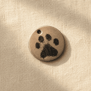 Round paw print earring charm in antique bronze finish with engraved black paw design, photographed on soft beige linen fabric in warm natural light.
