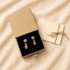 Gift-ready packaging photo featuring handmade paw print gemstone earrings displayed inside a kraft jewelry gift box with black velvet insert beside a tied message card on soft beige linen fabric.