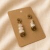Close-up product photograph of handmade paw print gemstone earrings displayed on a kraft jewelry card over soft beige linen fabric, featuring warm earthy gemstone tones and gold accents.
