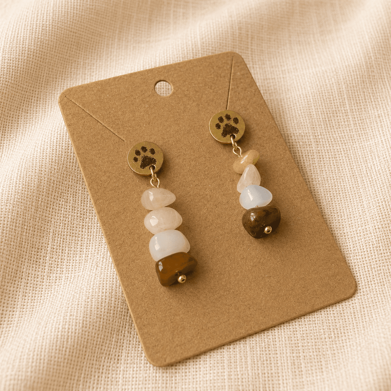 Close-up product photograph of handmade paw print gemstone earrings displayed on a kraft jewelry card over soft beige linen fabric, featuring warm earthy gemstone tones and gold accents.
