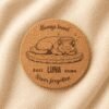 Top-down product photograph of a personalized round cork pet memorial coaster engraved with a sleeping cat illustration, the name Luna, memorial years 2011–2026, and the phrase “Never forgotten,” displayed on soft cream linen fabric.