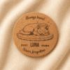 Top-down product photograph of a personalized round cork pet memorial coaster engraved with a sleeping cat illustration, the name Luna, memorial years 2011–2026, and the phrase “Never forgotten,” displayed on soft cream linen fabric.