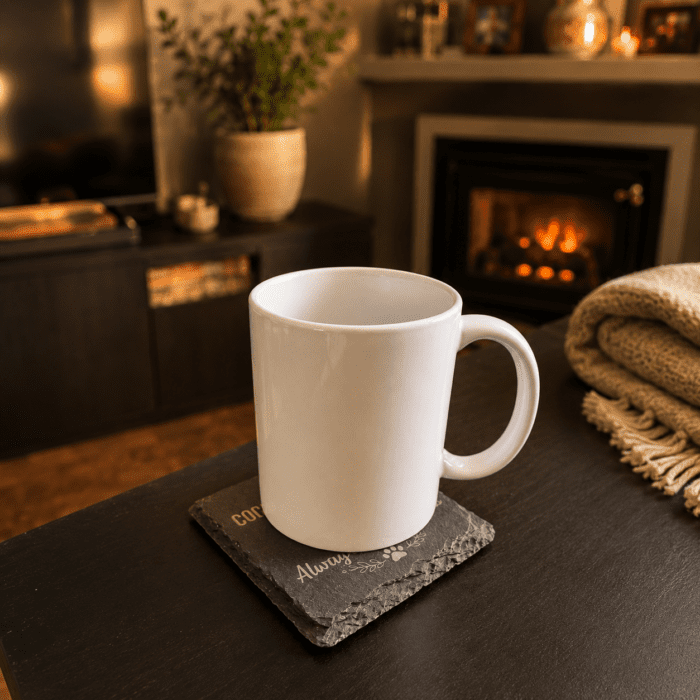 Warm lifestyle photograph of a white mug resting on a personalized slate memorial coaster beside a fireplace in a cozy living room setting.