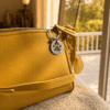 Warm lifestyle photo of a yellow handbag by a sunlit window with a personalized paw print memorial charm attached to the zipper, featuring the year “2011.”