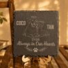 Personalized Pet Memorial Slate Coaster Warm lifestyle product photo of a personalized engraved slate memorial coaster displayed upright on a wooden easel, featuring a dog and cat illustration with the names “Coco” and “Tan” and the phrase “Always in Our Hearts.”