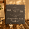 Warm lifestyle product photo of a personalized engraved slate memorial coaster displayed upright on a wooden easel, featuring a dog and cat illustration with the names “Coco” and “Tan” and the phrase “Always in Our Hearts.”