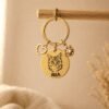 Gold personalized pet keychain hanging from a wooden rod, featuring an engraved cat portrait, the name “MIA,” a heart charm, and a paw charm in warm neutral lighting.