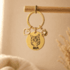 Gold personalized pet keychain hanging from a wooden rod, featuring an engraved cat portrait, the name “MIA,” a heart charm, and a paw charm in warm neutral lighting.